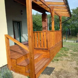 Drewniany taras z zadaszeniem z poliwęglanu, schodami i balustradą. Widok z boku na konstrukcję przylegającą do budynku z kominem w tle.