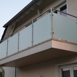 Carport Expert - Nowoczesna zabudowa balkonu z matowego szkła i metalowych słupków, widok z dołu, oświetlenie dzienne, fragment elewacji budynku w tle, minimalistyczny design.
