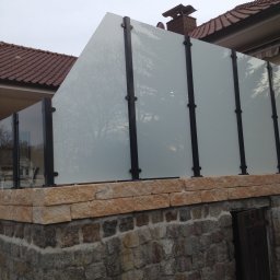 Carport Expert - Nowoczesne ogrodzenie z matowego szkła i czarnych słupków, osadzone na kamiennym murku. Widok na dach budynku w tle. Minimalistyczny design.