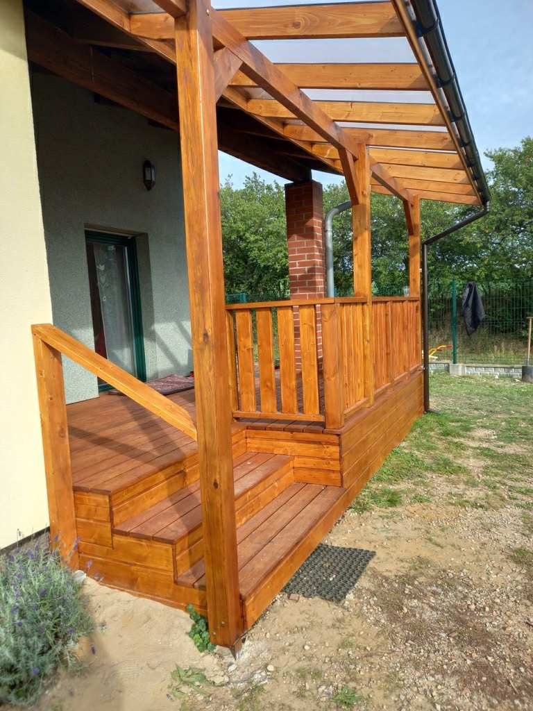 Drewniany taras z zadaszeniem z poliwęglanu, schodami i balustradą. Widok z boku na konstrukcję przylegającą do budynku z kominem w tle.