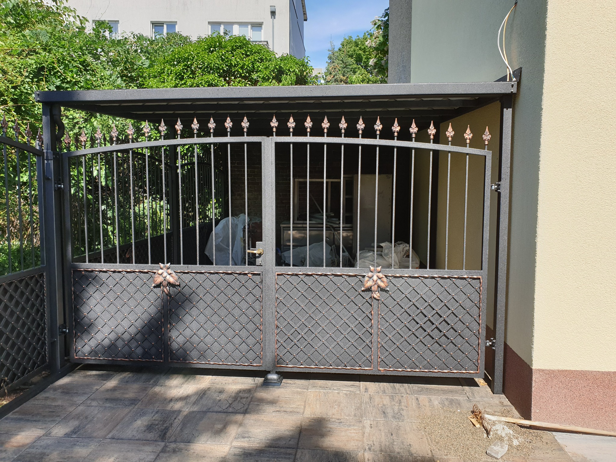 Metalowa brama wjazdowa z ozdobnymi elementami pod zadaszeniem, widok z zewnątrz. Brama w kolorze grafitowym z miedzianymi akcentami.