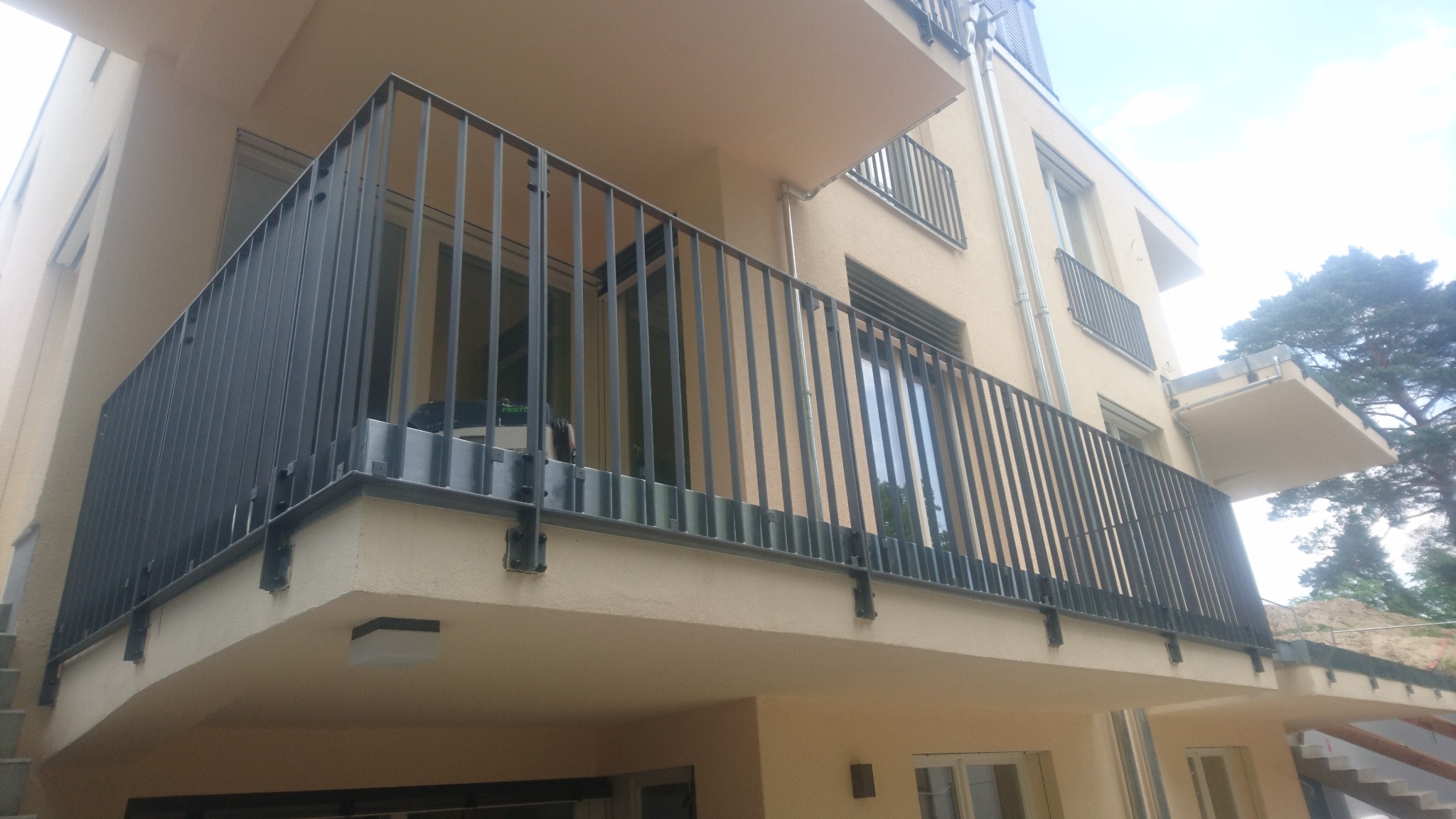 Nowoczesna balustrada balkonowa z pionowych, czarnych prętów, zamocowana do beżowej elewacji budynku. Widok z dołu, podkreślający minimalistyczny design i solidne wykonanie.