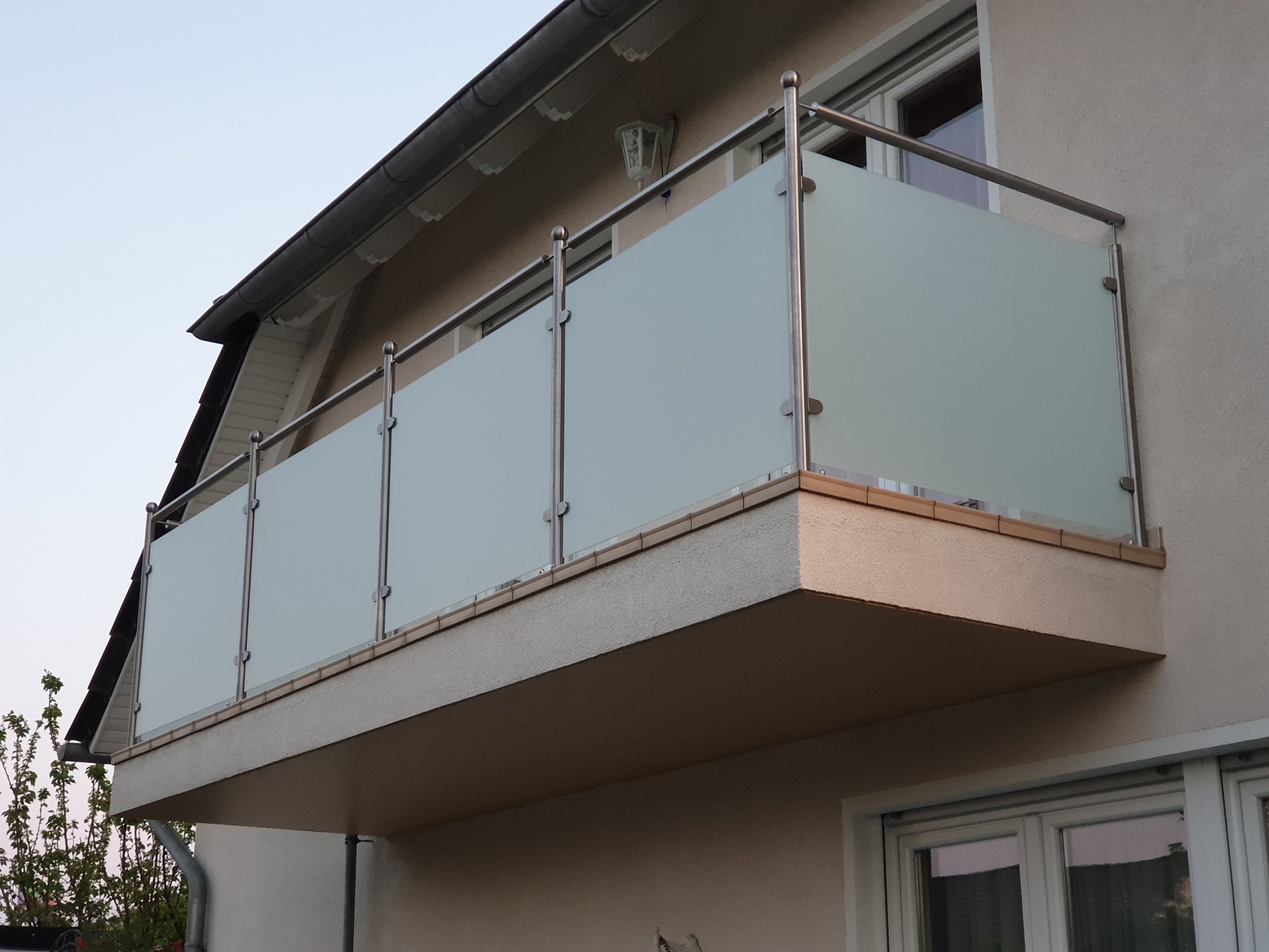 Nowoczesna zabudowa balkonu z matowego szkła i metalowych słupków, widok z dołu, oświetlenie dzienne, fragment elewacji budynku w tle, minimalistyczny design.