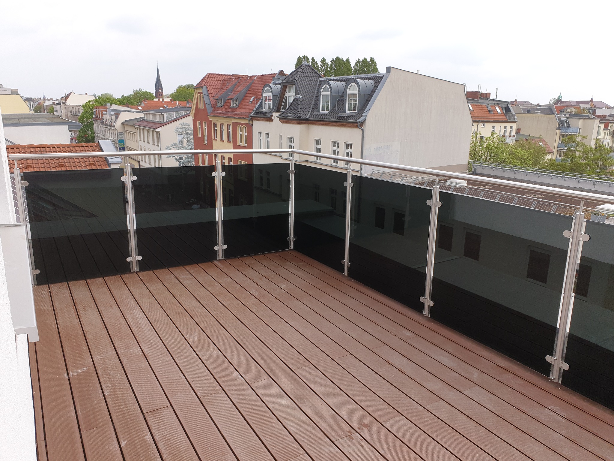 Nowoczesny balkon z brązową podłogą z desek i szklanymi panelami balustrady ze stali nierdzewnej. Widok na budynki mieszkalne w tle.