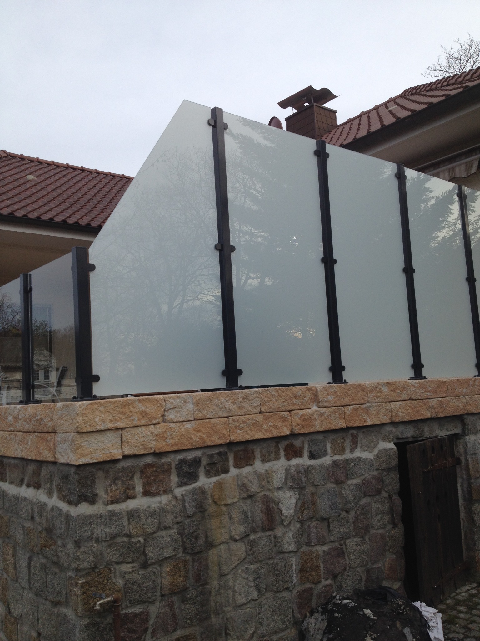 Nowoczesne ogrodzenie z matowego szkła i czarnych słupków, osadzone na kamiennym murku. Widok na dach budynku w tle. Minimalistyczny design.