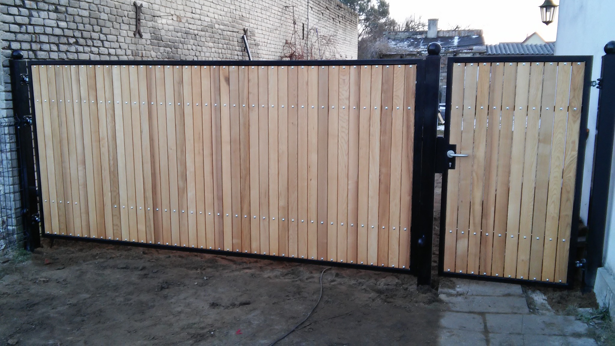 Nowoczesna brama z jasnego drewna i czarnej metalowej ramy, widoczna z bliska, z wyraźnymi detalami montażu i fakturą drewna. Brama dwuskrzydłowa z furtką.