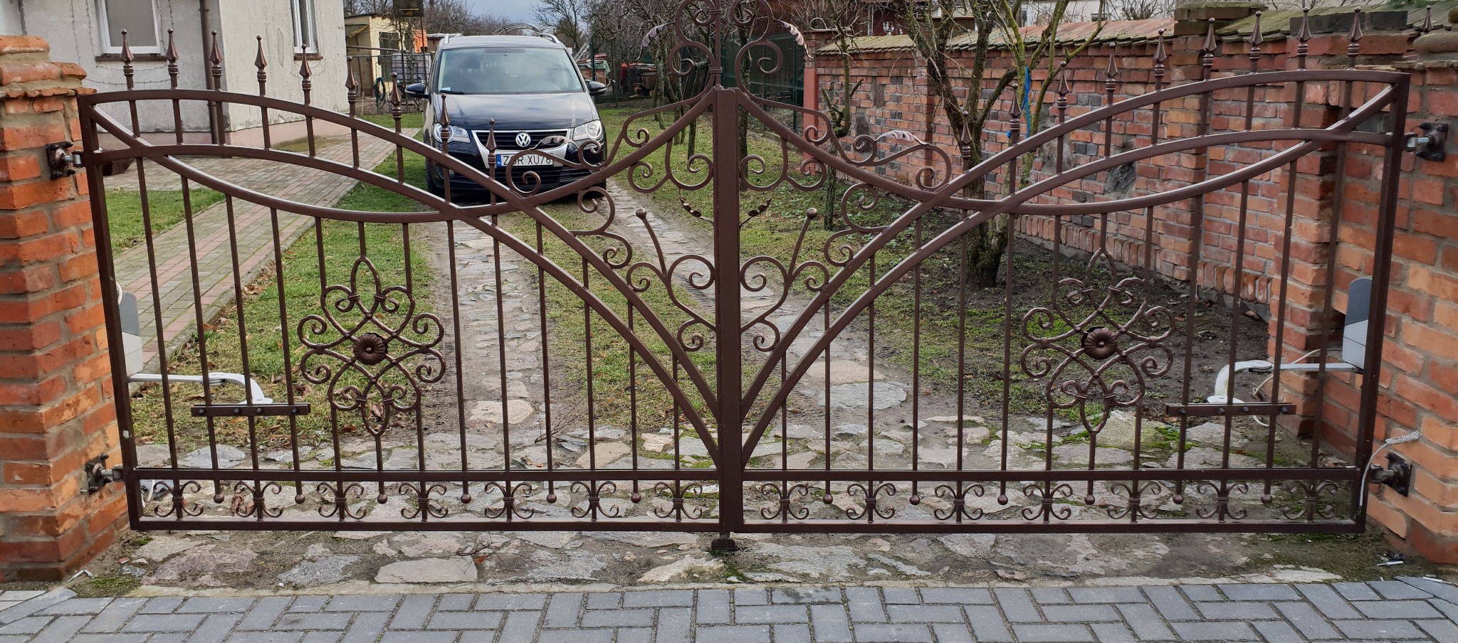 Kuta brama wjazdowa z automatyką, zdobiona ornamentami, zamykająca wjazd na posesję z widocznym samochodem i ceglanym murem w tle. Brązowa kolorystyka.