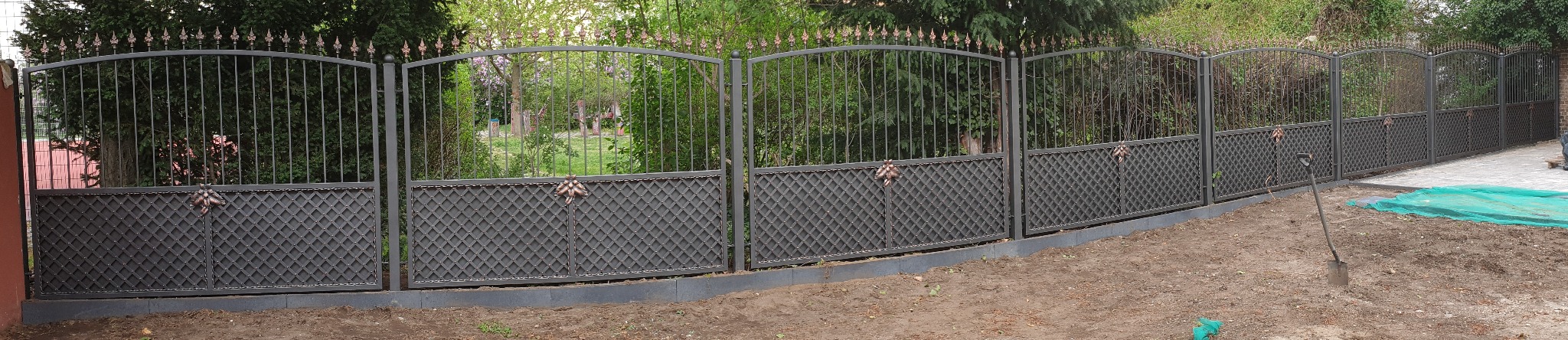 Długi, metalowy płot z ozdobnymi grotami i romboidalnym wypełnieniem dolnej części, otoczony zielenią ogrodu. Widoczna łopata wbita w ziemię.