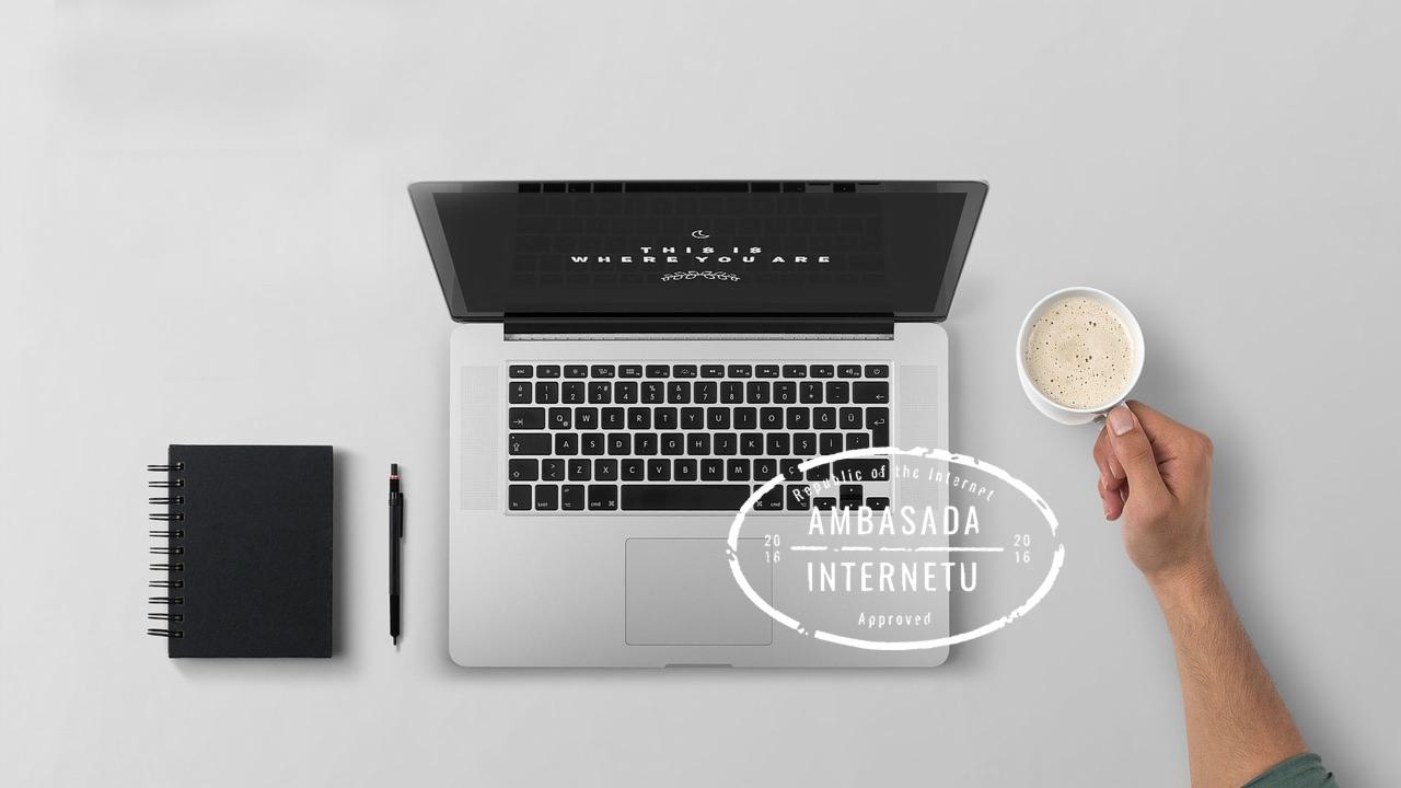 Widok z góry na biurko z laptopem z napisem 'This is where you are' na ekranie, notatnikiem, długopisem i ręką trzymającą filiżankę kawy, z nałożonym stemplem 'Ambasada Internetu'.