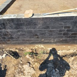 Jar-bud - Czarne, zaizolowane ściany fundamentowe z widocznymi numerami '320' i '078', z łopatą i kartonowym pudełkiem na górze, oraz cień osoby na ziemi.