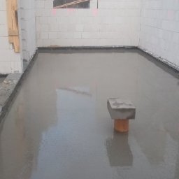 Jar-bud - Świeżo wylana, gładka posadzka betonowa w surowym wnętrzu budynku z białych bloczków, widoczny pion kanalizacyjny wystający z podłogi, na zewnątrz budynku widoczny teren budowy.