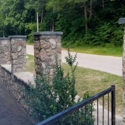 Jar-bud - Kamienne słupki ogrodzeniowe z betonowymi nakryciami, tworzące mur wzdłuż drogi, widoczne spod czarnego metalowego ogrodzenia.