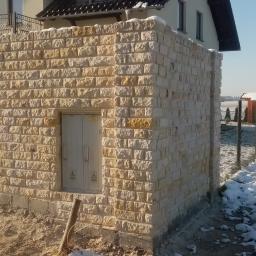 Jar-bud - Murowane ogrodzenie z jasnego kamienia łamanego, z wbudowanymi drzwiczkami technicznymi, na tle zimowego krajobrazu z domem w oddali.