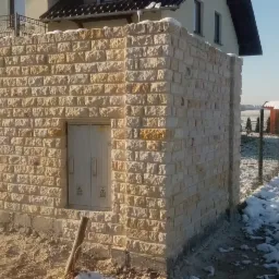 Murowane ogrodzenie z jasnego kamienia łamanego, z wbudowanymi drzwiczkami technicznymi, na tle zimowego krajobrazu z domem w oddali.