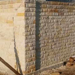 Jar-bud - Wzniesiony fragment ogrodzenia z kamienia naturalnego, wykonanego z małych, regularnych bloczków w odcieniach beżu i brązu, widoczny element drewnianego stelażu pomocniczego i paleta z resztkami...