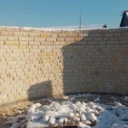 Jar-bud - Niedokończona, zakrzywiona ściana z jasnego kamienia, częściowo pokryta śniegiem, w zimowej scenerii z widocznymi budynkami w tle.