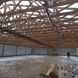 Wnętrze budowanego budynku z drewnianą konstrukcją dachu, widoczne ściany z betonowych bloczków i dwóch pracowników w oddali.