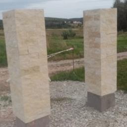 Jar-bud - Dwa kamienne słupki ogrodzeniowe z jasnego piaskowca na betonowej podstawie, teren wokół wysypany drobnym kamieniem, w tle polna droga i zieleń.
