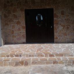 Jar-bud - Kamienny podest wejściowy z trzema stopniami, prowadzący do ciemnych drzwi z owalnym przeszkleniem, otoczonych ścianą z nieregularnego kamienia w odcieniach beżu i brązu.