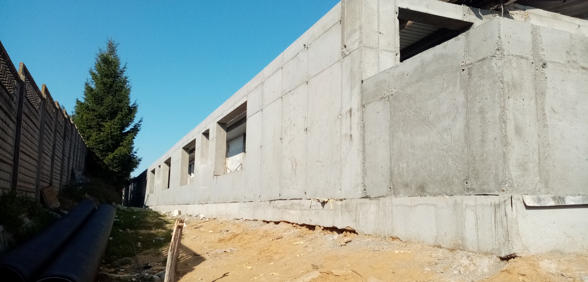 Surowy budynek żelbetowy w trakcie budowy z widocznymi otworami okiennymi, obok ogrodzenie i drzewo iglaste na tle błękitnego nieba.