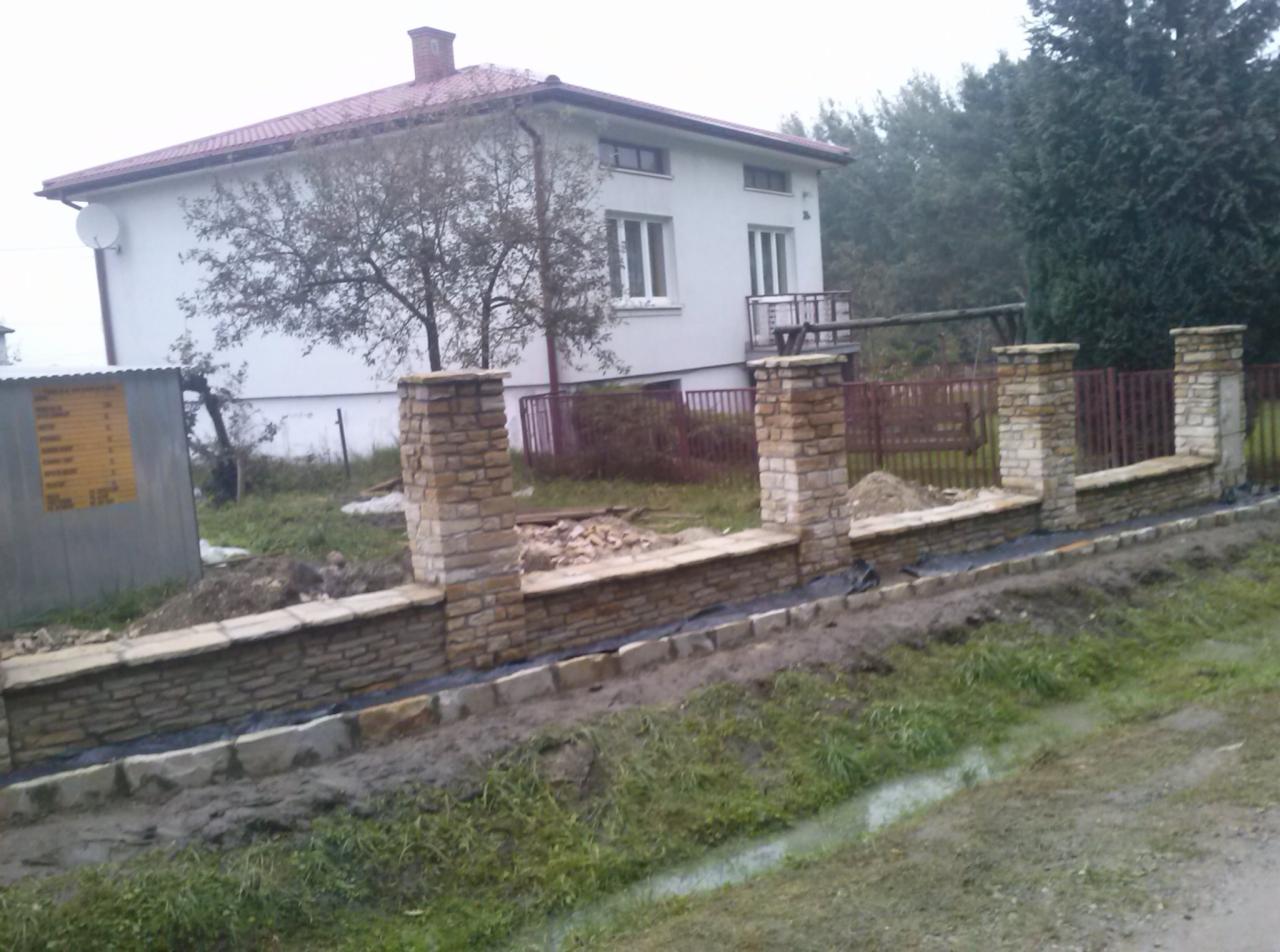 Wznoszenie kamiennego murku ogrodzeniowego z filarami z jasnego kamienia, widoczny etap budowy z czarną folią izolacyjną, w tle dom z czerwonym dachem i metalowe ogrodzenie.