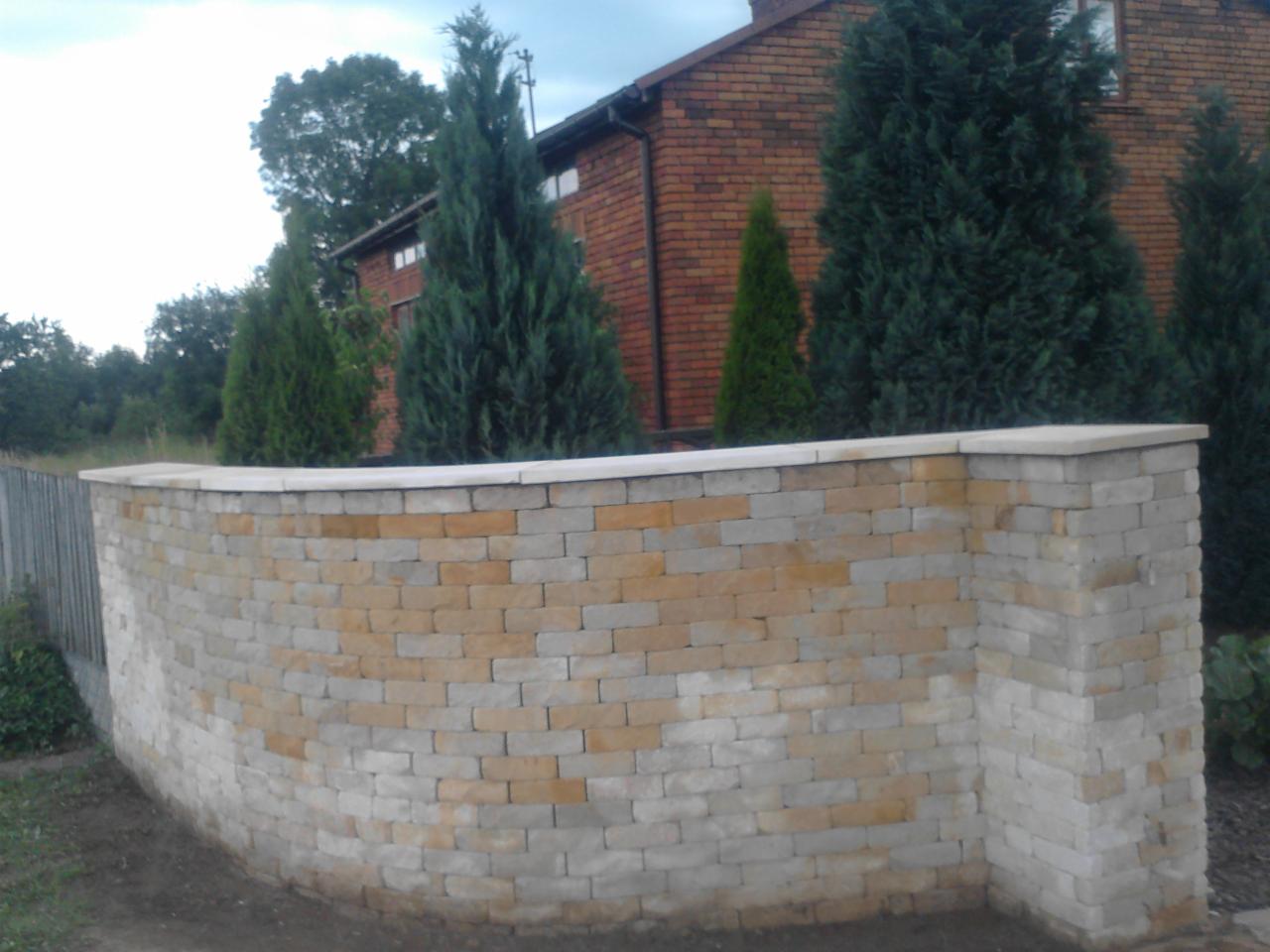 Murek oporowy z piaskowca o zaokrąglonym kształcie, zwieńczony jasnymi płytami, na tle ceglanego domu i iglaków.