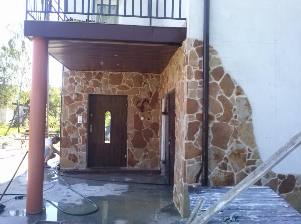 Wykończenie elewacji wejścia do domu kamieniem naturalnym, widoczny fragment balkonu, kolumna podtrzymująca zadaszenie i pracownik czyszczący powierzchnię po zakończeniu prac.