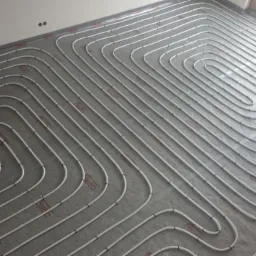 Białe rurki ogrzewania podłogowego ułożone spiralnie na szarej macie izolacyjnej w jasnym pomieszczeniu z oknem.