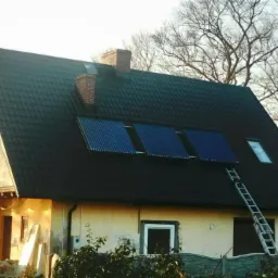 Dwukondygnacyjny dom z ciemną dachówką, na której zamontowano trzy niebieskie kolektory słoneczne; drabina oparta o ścianę, widoczne drzewa bez liści w tle.