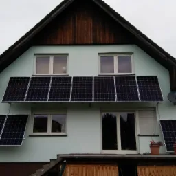 Dom z zainstalowanymi panelami słonecznymi na elewacji, widoczne okna i satelita na ścianie, dwa kwiaty w doniczkach na balkonie.