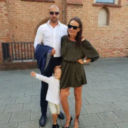 Elegancka rodzina pozuje na tle ceglanej ściany kościoła. Mężczyzna trzyma marynarkę, kobieta w sukience i szpilkach, a dziecko w białej koszuli i krótkich spodenkach.