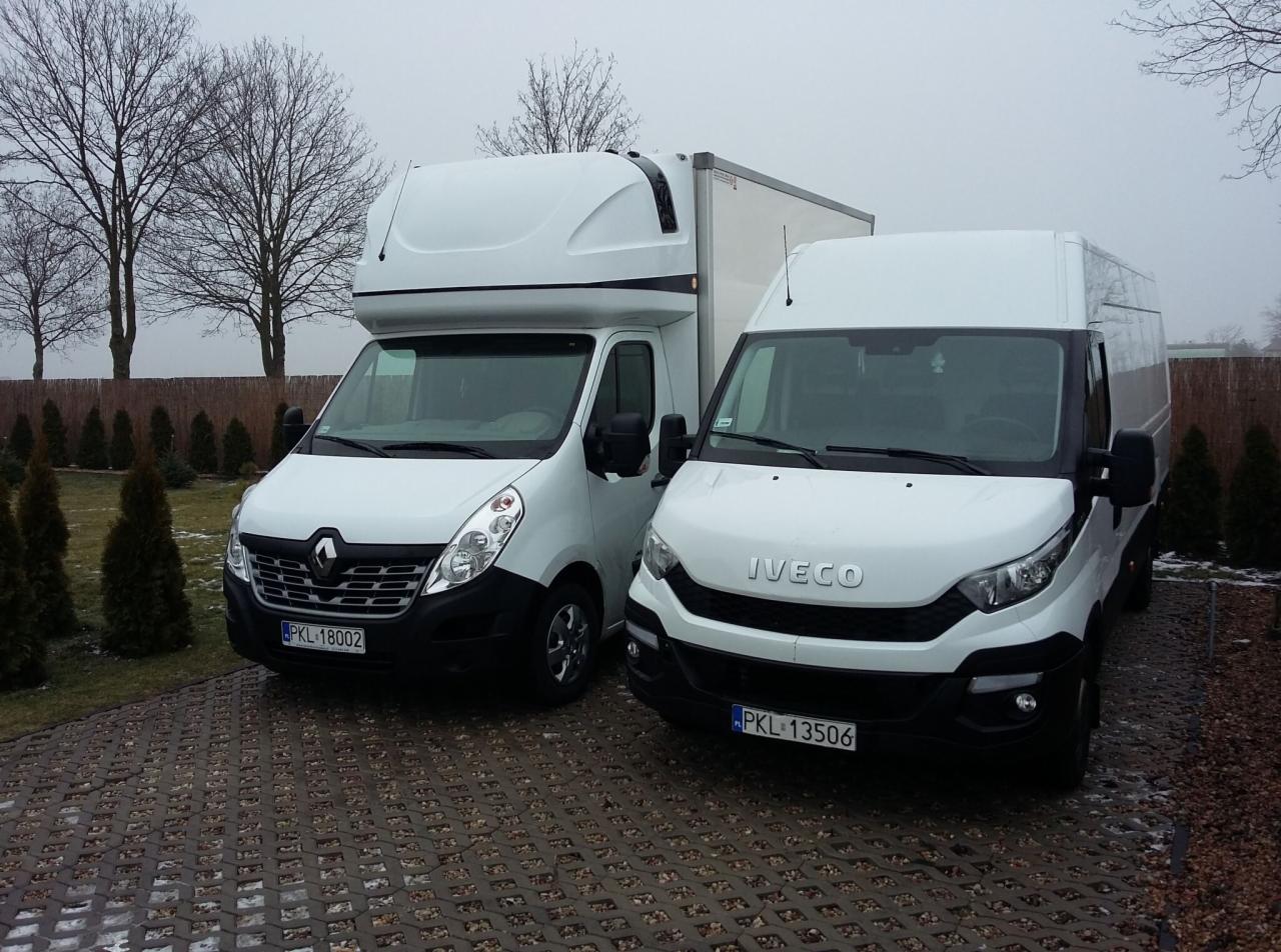 Dwa białe busy dostawcze, Renault Master z zabudową kontenerową i Iveco Daily, zaparkowane na kostce brukowej w pochmurny dzień, widoczne tablice rejestracyjne.
