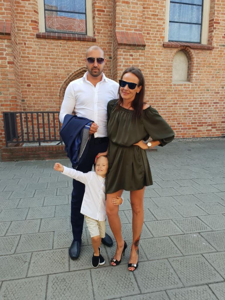 Elegancka rodzina pozuje na tle ceglanej ściany kościoła. Mężczyzna trzyma marynarkę, kobieta w sukience i szpilkach, a dziecko w białej koszuli i krótkich spodenkach.