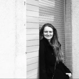 Portret kobiety w ciemnym płaszczu, opierającej się o ścianę z żaluzjami, uśmiechającej się do kamery w czarno-białej tonacji.