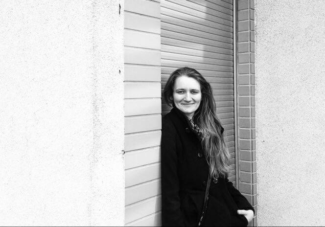 Portret kobiety w ciemnym płaszczu, opierającej się o ścianę z żaluzjami, uśmiechającej się do kamery w czarno-białej tonacji.