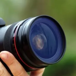 Czarny aparat fotograficzny z dużym obiektywem trzymany w dłoniach na rozmytym zielonym tle.