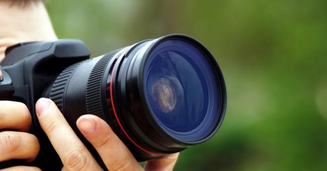 Czarny aparat fotograficzny z dużym obiektywem trzymany w dłoniach na rozmytym zielonym tle.