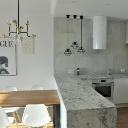 Nowoczesna kuchnia w odcieniach bieli i szarości z marmurowym blatem, drewnianym stołem i designerskimi lampami wiszącymi oraz plakatem Vogue na ścianie.