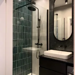 Nowoczesna łazienka z kabiną prysznicową typu walk-in, ciemnozielonymi płytkami ułożonymi pionowo, czarną armaturą i owalnym lustrem z czarną ramą nad białą umywalką nablatową na czarnej szafce.