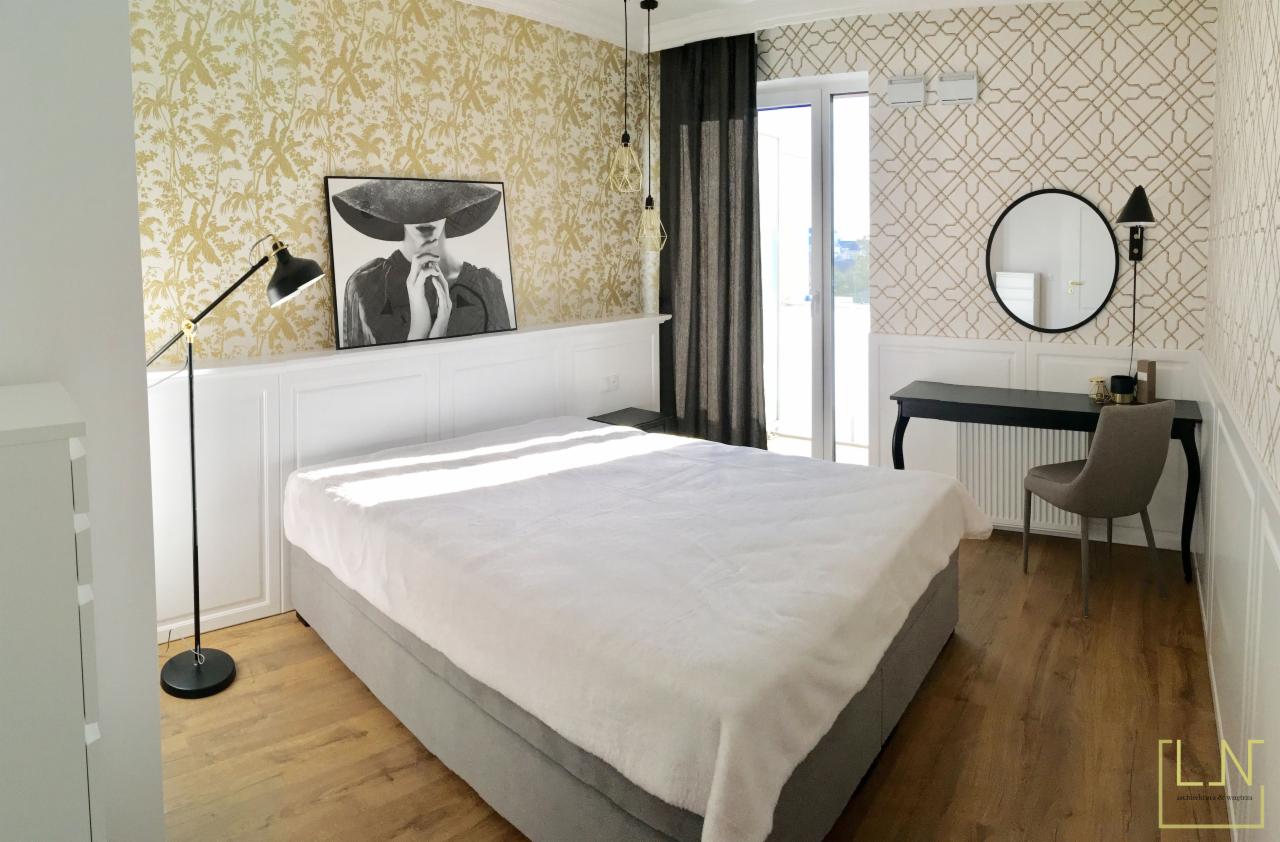 Jasna sypialnia z łóżkiem kontynentalnym, drewnianą podłogą, tapetą w złote wzory i czarno-białym portretem kobiety w kapeluszu nad wezgłowiem, uzupełniona toaletką z okrągłym lustrem.