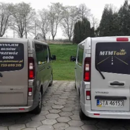 Dwa busy Opel Vivaro, jeden beżowy, drugi srebrny, zaparkowane tyłem z naklejkami reklamowymi firmy MTM Tour oferującej wynajem busów i przewóz osób, widoczne numery rejestracyjne i zieleń w tle.