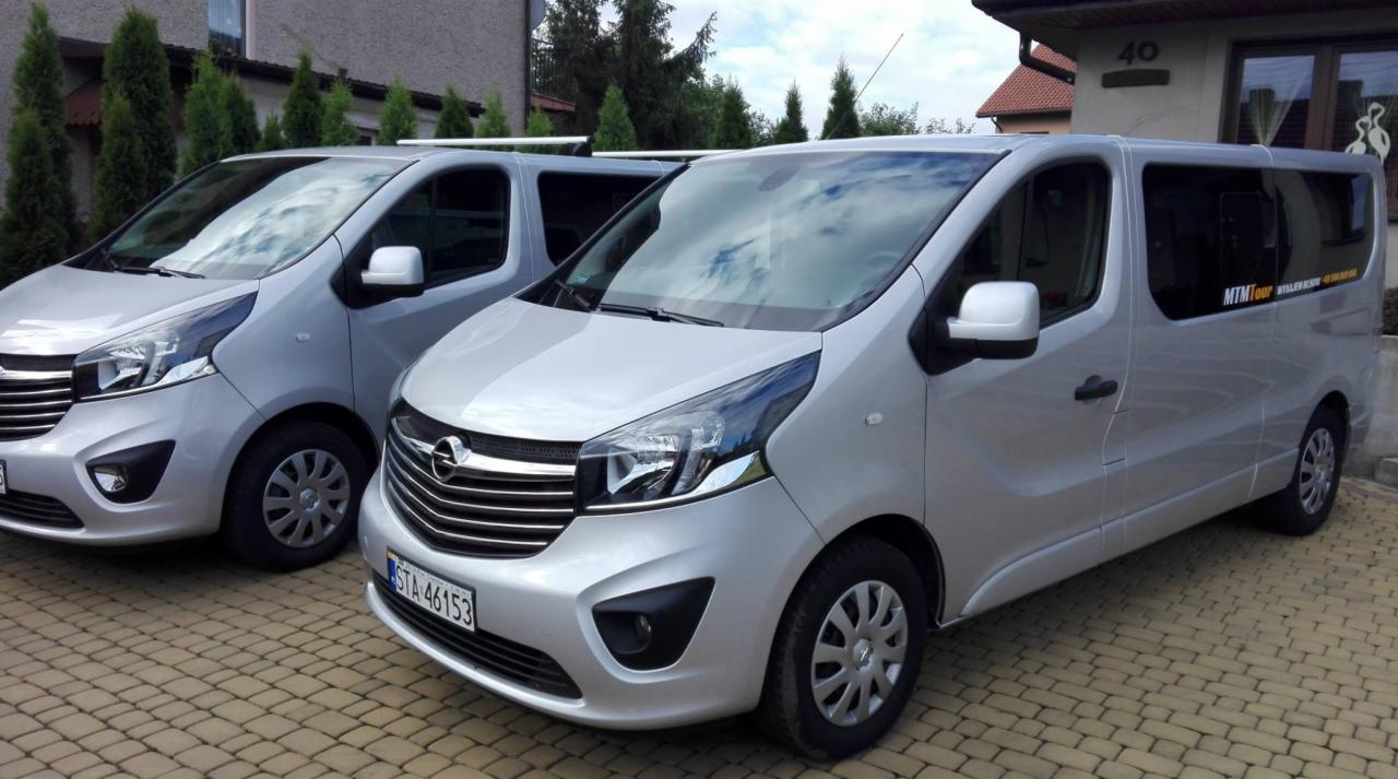 Dwa srebrne busy Opel Vivaro zaparkowane na brukowanym podjeździe przed domem z numerem 40, widoczne logo firmy MTM Tour na przyciemnianych szybach.