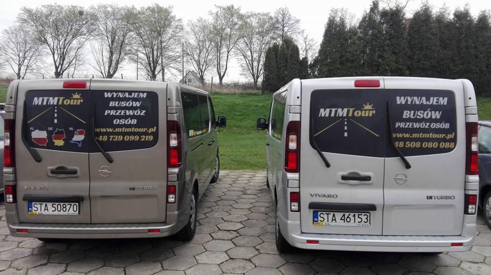 Dwa busy Opel Vivaro, jeden beżowy, drugi srebrny, zaparkowane tyłem z naklejkami reklamowymi firmy MTM Tour oferującej wynajem busów i przewóz osób, widoczne numery rejestracyjne i zieleń w tle.