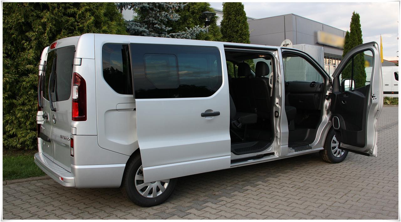 Srebrny van z otwartymi przesuwnymi drzwiami i drzwiami kierowcy, zaparkowany na kostce brukowej przed budynkiem i zielonymi drzewami.