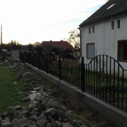 Nowo postawione, czarne, metalowe ogrodzenie z ozdobnymi elementami na betonowej podmurówce, widok z boku, w tle biały dom z brązowymi oknami i fragment drogi.