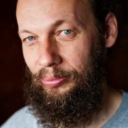 Portret brodatego mężczyzny z jasnymi oczami, uchwycony w naturalnym świetle, z subtelnym uśmiechem i widocznymi zmarszczkami wokół oczu.