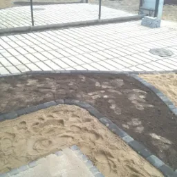 Układanie nawierzchni z ażurowych płyt betonowych i obrzeży chodnikowych, widoczne warstwy piasku i ziemi.