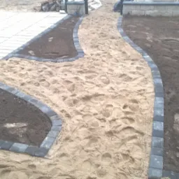 Kształtowana ścieżka z piasku i obrzeży z kostki brukowej, fragment trawnika z świeżo nawiezioną ziemią, w tle ułożona kostka brukowa.