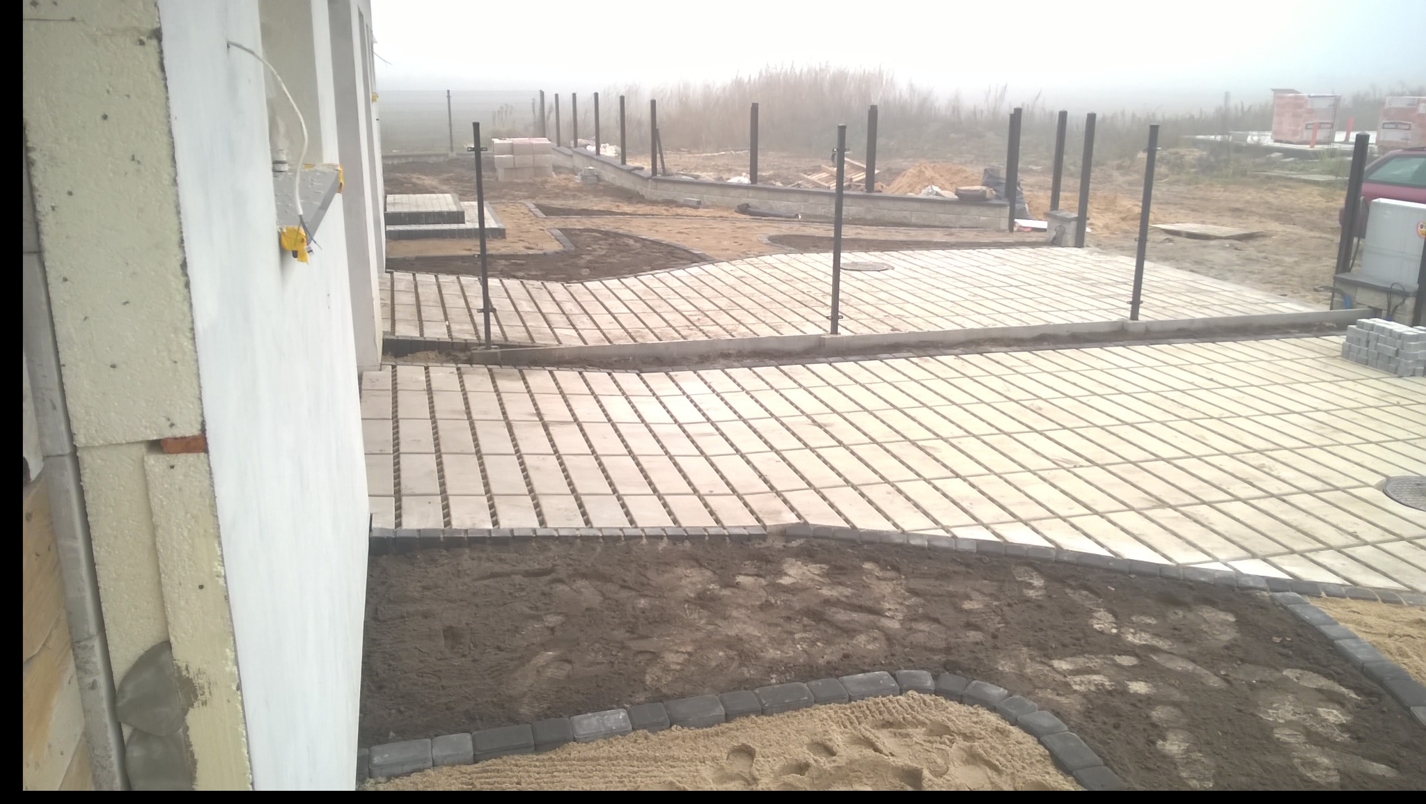 Układanie kostki brukowej na posesji w trakcie budowy, widoczne fundamenty domu i przygotowane podłoże pod ogród, mglista pogoda.