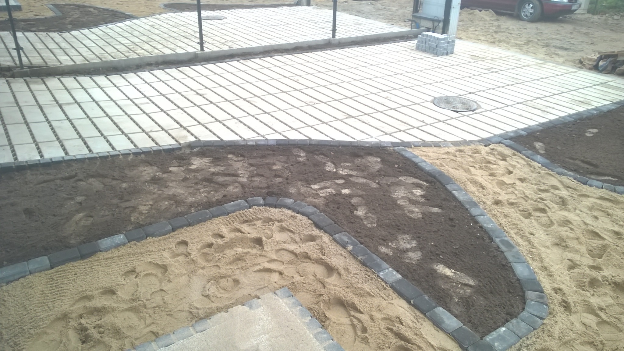 Układanie nawierzchni z ażurowych płyt betonowych i obrzeży chodnikowych, widoczne warstwy piasku i ziemi.
